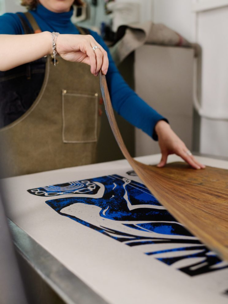 Cropped shot of young female artisan standing by printing machine and taking top sheet off paper with blue sketch or artwork
